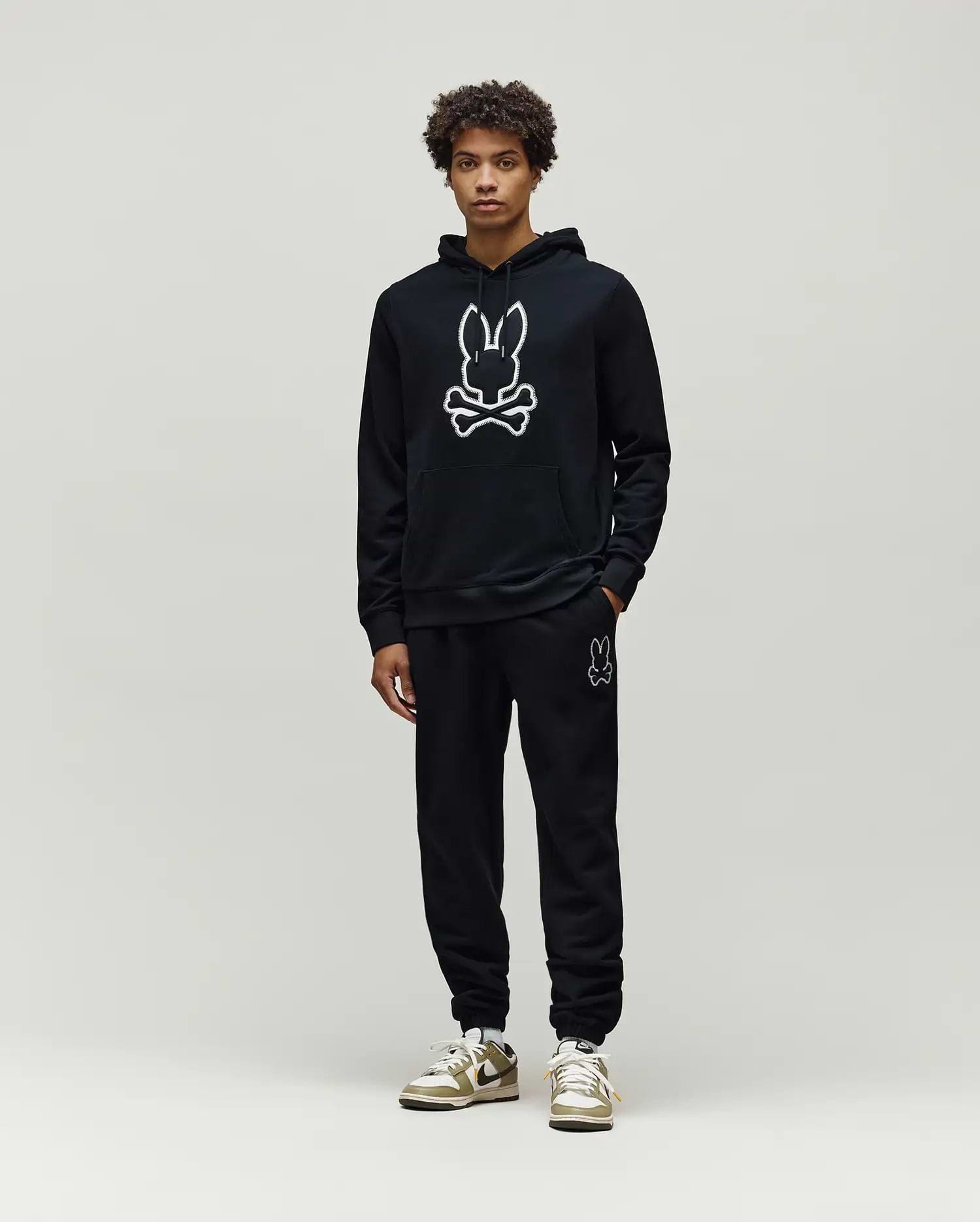 Mens Gage Patch Hoodie: Black relaxed fit French terry hoodie with a large white outlined Psycho Bunny skull logo on the chest, kangaroo pocket, and ribbed cuffs, paired with matching black sweatpants.
