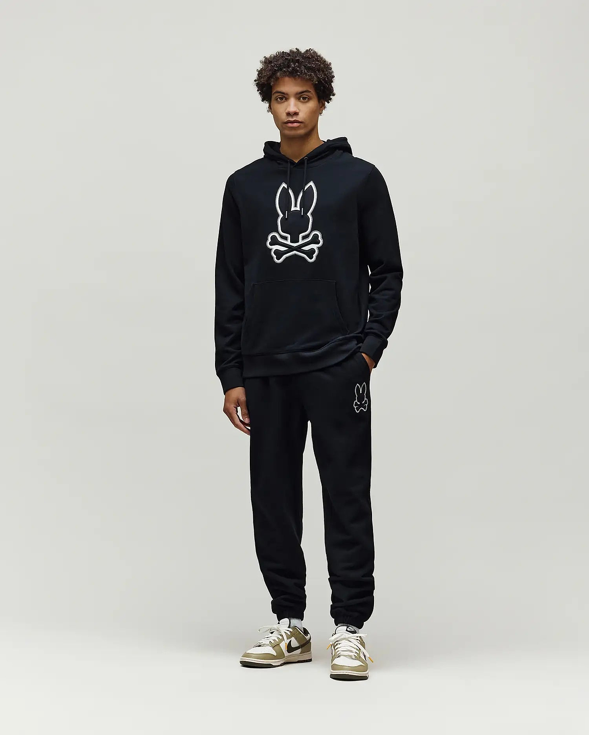 Mens Gage Patch Hoodie: Black relaxed fit French terry hoodie with a large white outlined Psycho Bunny skull logo on the chest, kangaroo pocket, and ribbed cuffs, paired with matching black sweatpants.