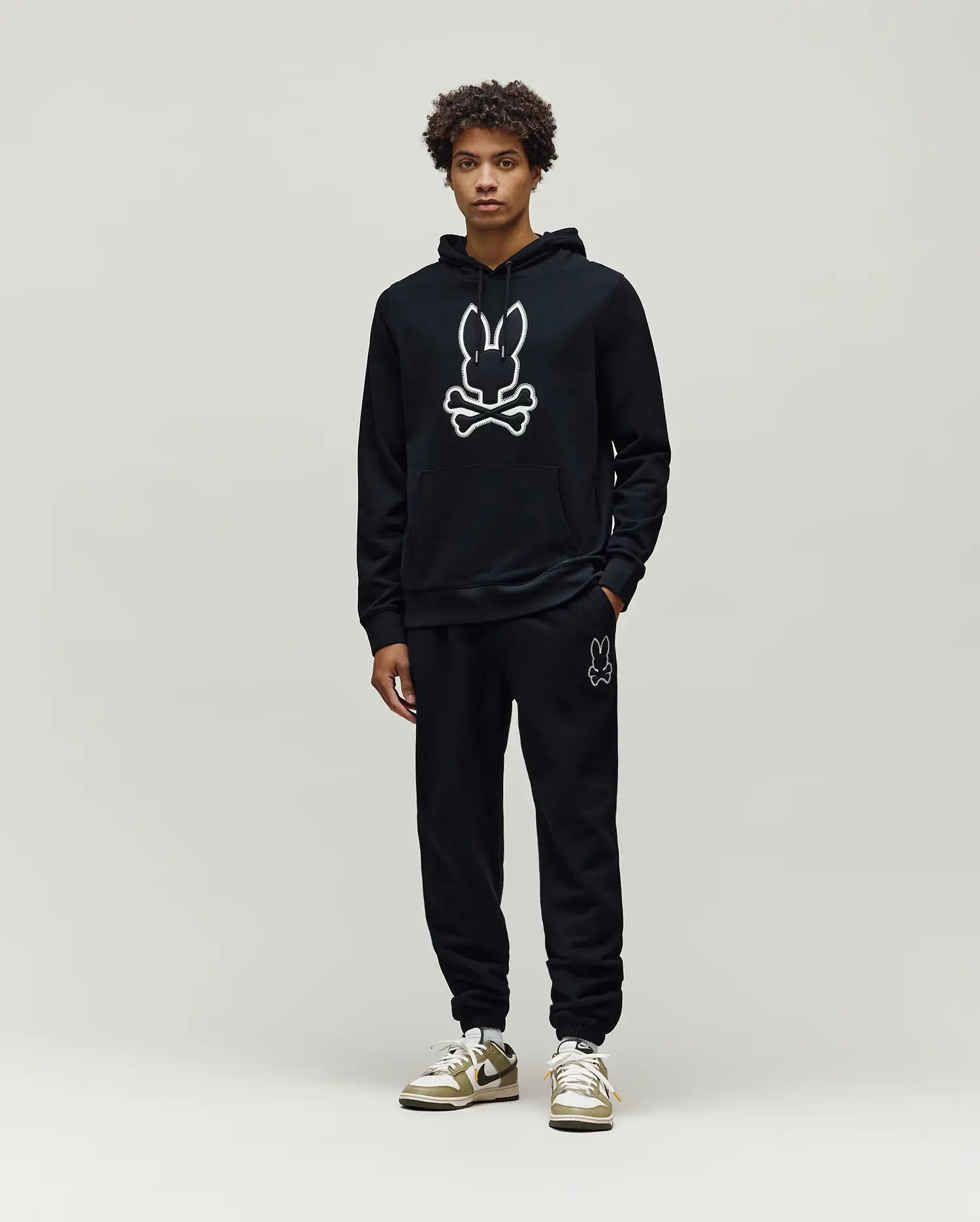 Mens Gage Patch Hoodie: Black relaxed fit French terry hoodie with a large white outlined Psycho Bunny skull logo on the chest, kangaroo pocket, and ribbed cuffs, paired with matching black sweatpants.