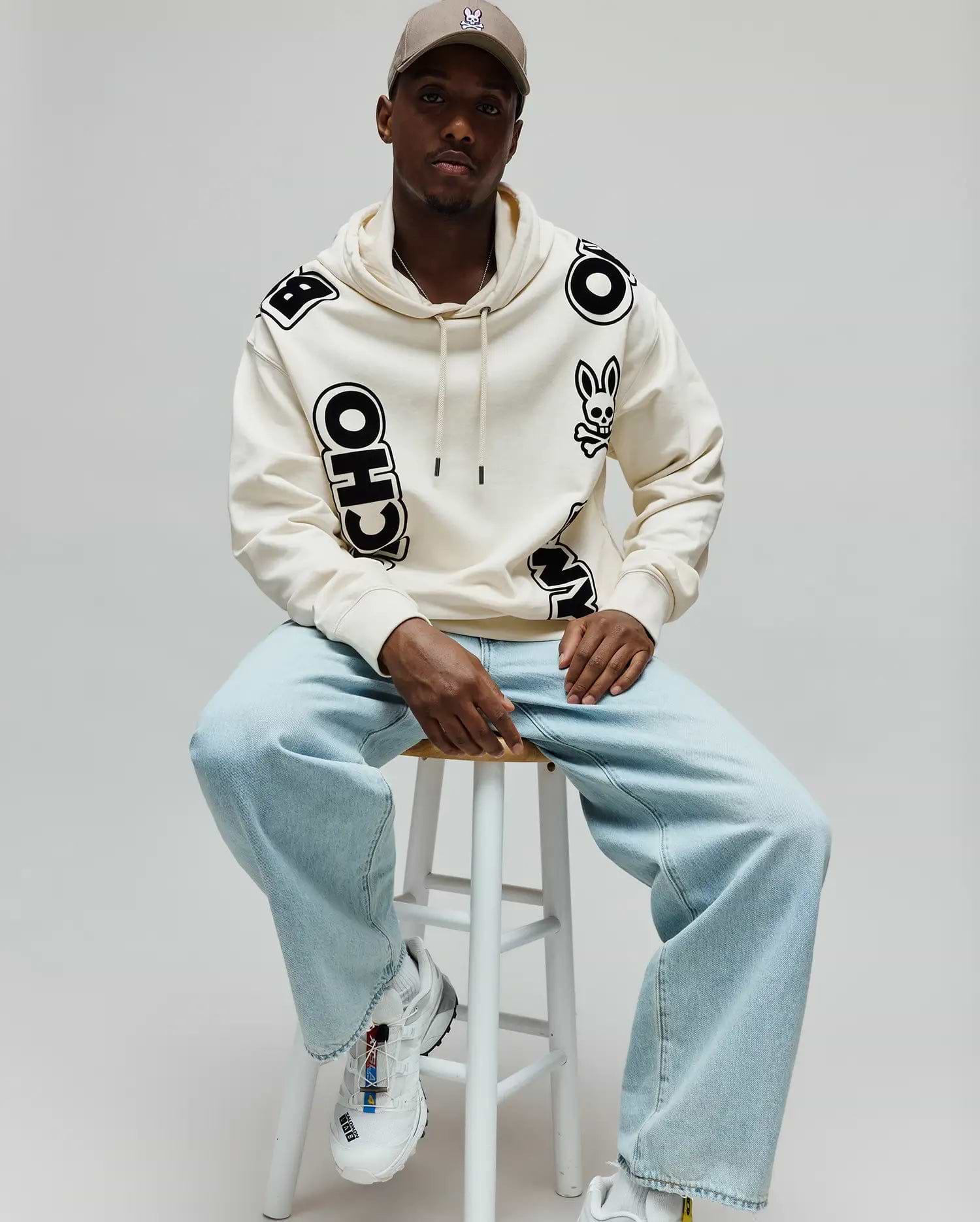 Mens Toby Oversized Hoodie in cream, featuring bold black scattered PSYCHO BUNNY lettering and a prominent bunny skull logo on the left chest, made from soft fleece.