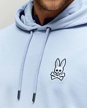 Mens Timothy 4k Bunny Hoodie, a light blue classic fit jersey hoodie with a small embroidered white and navy Psycho Bunny skull logo on the left chest and branded silver-tipped drawstrings.