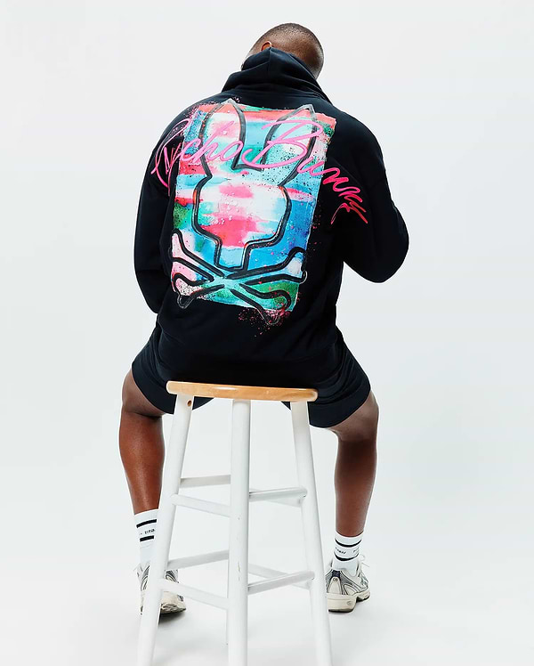 Mens Isaac Print Hoodie, black relaxed fit French terry hoodie with a large, vibrant paint-splatter Psycho Bunny skull graphic in blue, pink, green, and white, featuring neon pink script on the back.