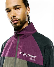 Mens Alder Nylon Colorblock Jacket in purple, dark grey, olive, with crinkled texture, full-zip, high collar, and white Psycho Bunny text logo with Designed For Disruption and coordinates on chest.