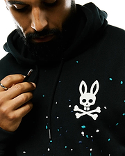Mens Malcom Splatter Hoodie, a black relaxed fit French terry hoodie with a large white embroidered Psycho Bunny skull and crossbones logo on the chest, accented by an all-over white, teal, and navy splatter print.