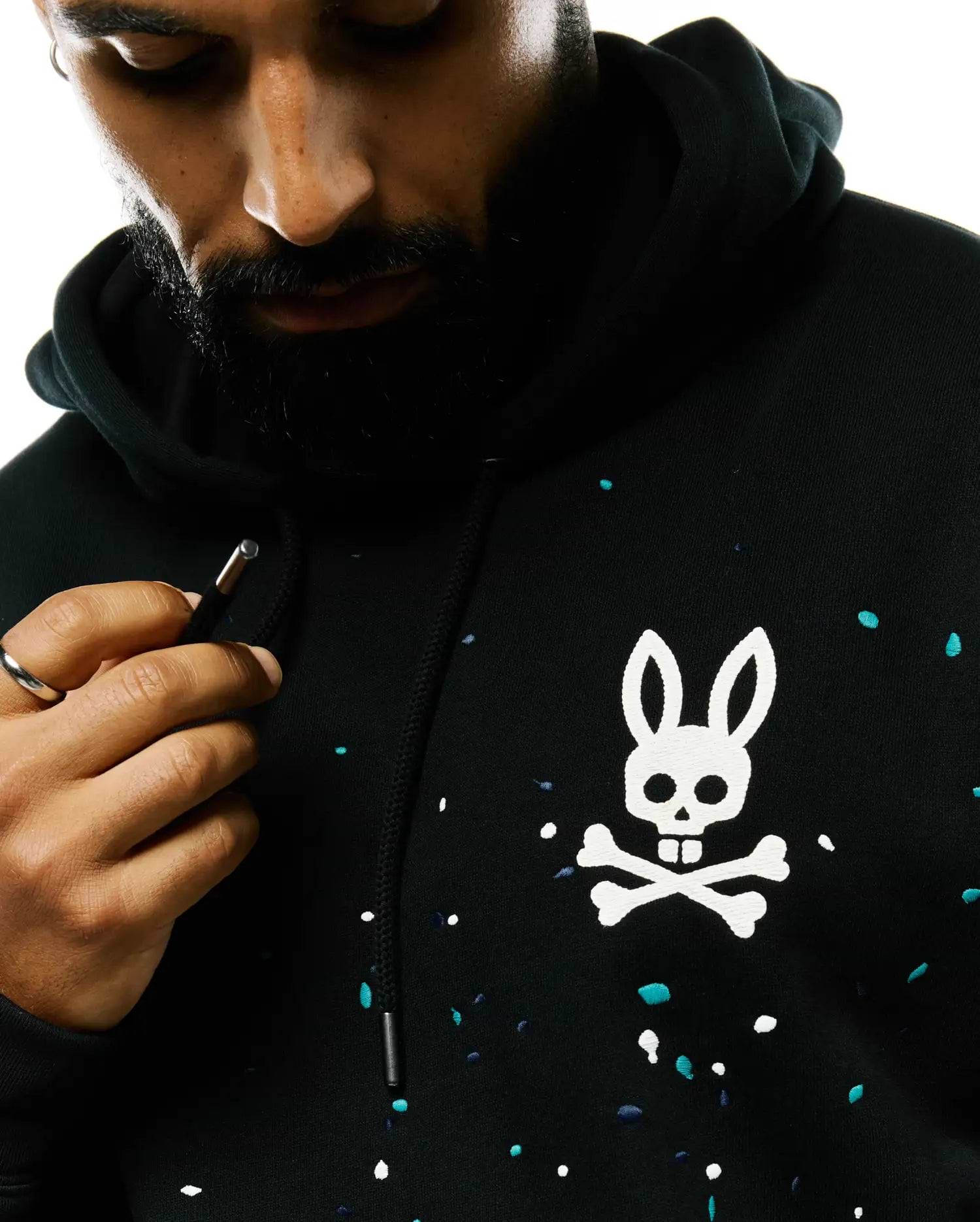Mens Malcom Splatter Hoodie, a black relaxed fit French terry hoodie with a large white embroidered Psycho Bunny skull and crossbones logo on the chest, accented by an all-over white, teal, and navy splatter print.