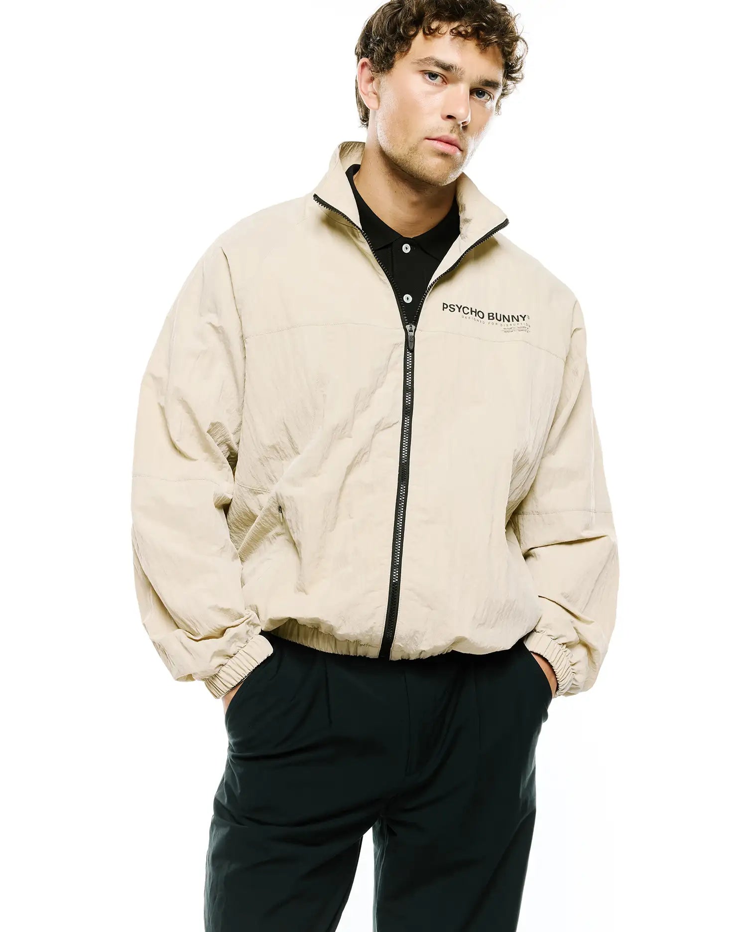 Mens Alder Nylon Jacket, a relaxed fit beige crinkled nylon full-zip jacket with a stand collar, elastic cuffs, and a black Psycho Bunny text logo with Designed For Disruption on the left chest.
