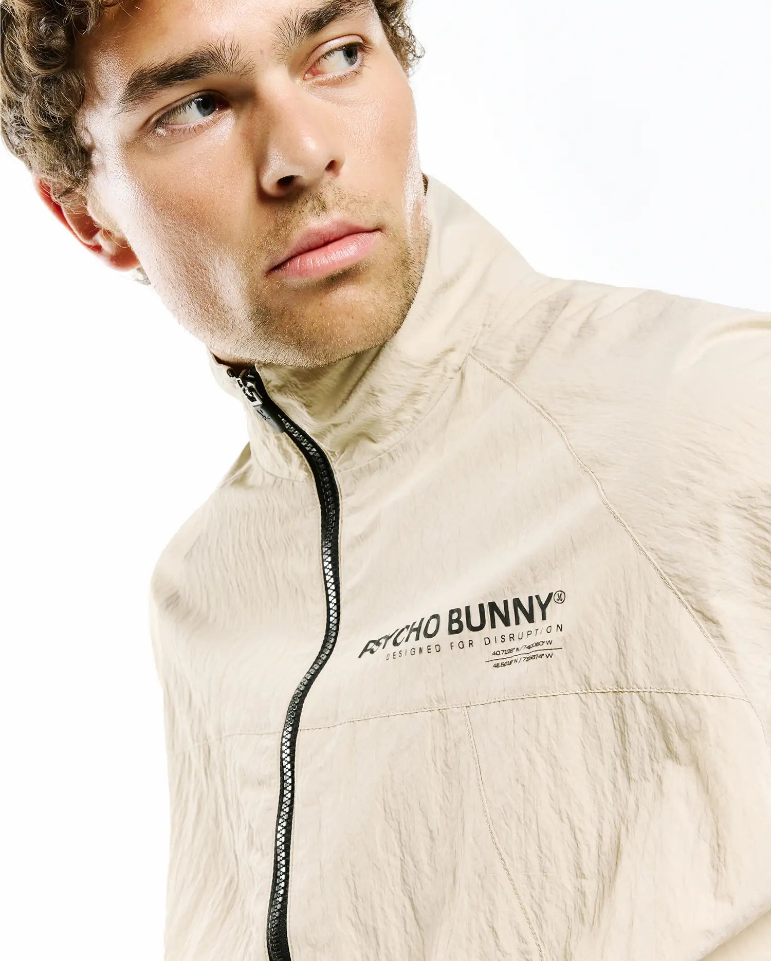Mens Alder Nylon Jacket in beige crinkled nylon, featuring a classic fit, high collar, black full-zip, and a black Psycho Bunny Designed For Disruption text logo with coordinates on the left chest.