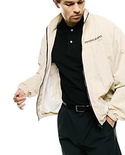 Mens Alder Nylon Jacket, a relaxed fit beige crinkled nylon full-zip jacket with black zipper and a black PSYCHO BUNNY DESIGNED FOR DISRUPTION text logo on left chest, over a black pique polo.