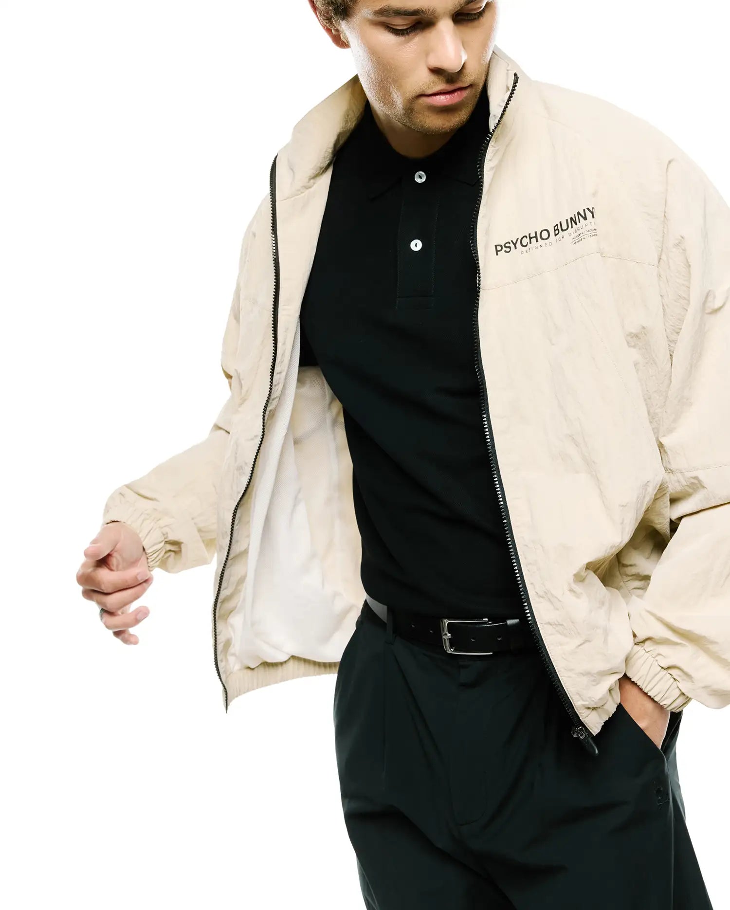 Mens Alder Nylon Jacket, a relaxed fit beige crinkled nylon full-zip jacket with black zipper and a black PSYCHO BUNNY DESIGNED FOR DISRUPTION text logo on left chest, over a black pique polo.