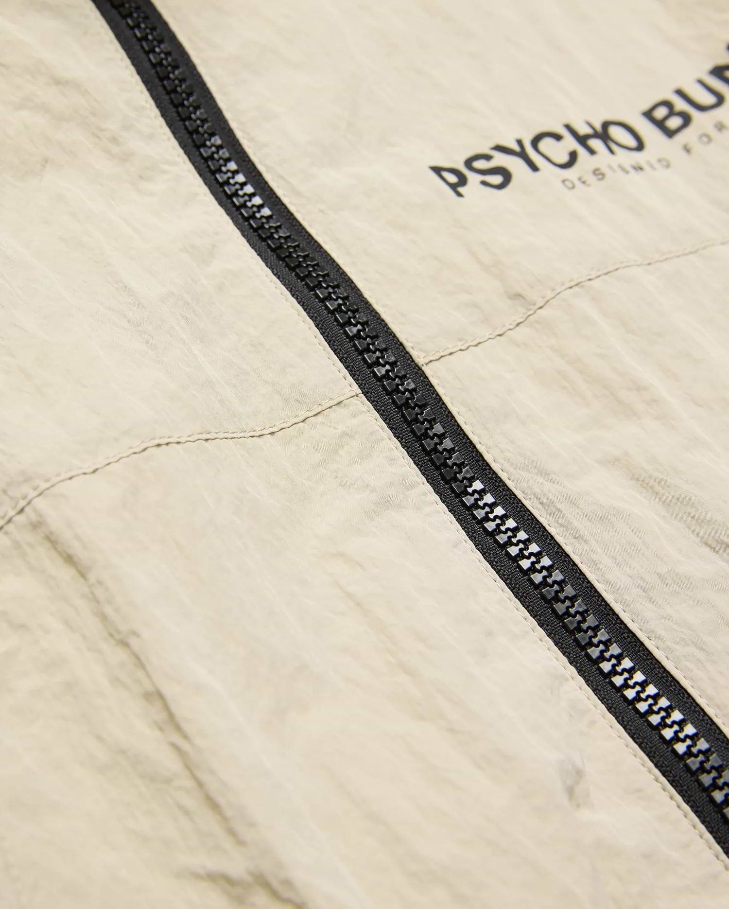Mens Alder Nylon Jacket, a relaxed fit light beige crinkled nylon windbreaker featuring a prominent black full-zip closure and bold black PSYCHO BUNNY DESIGNED FOR text print on the upper chest.