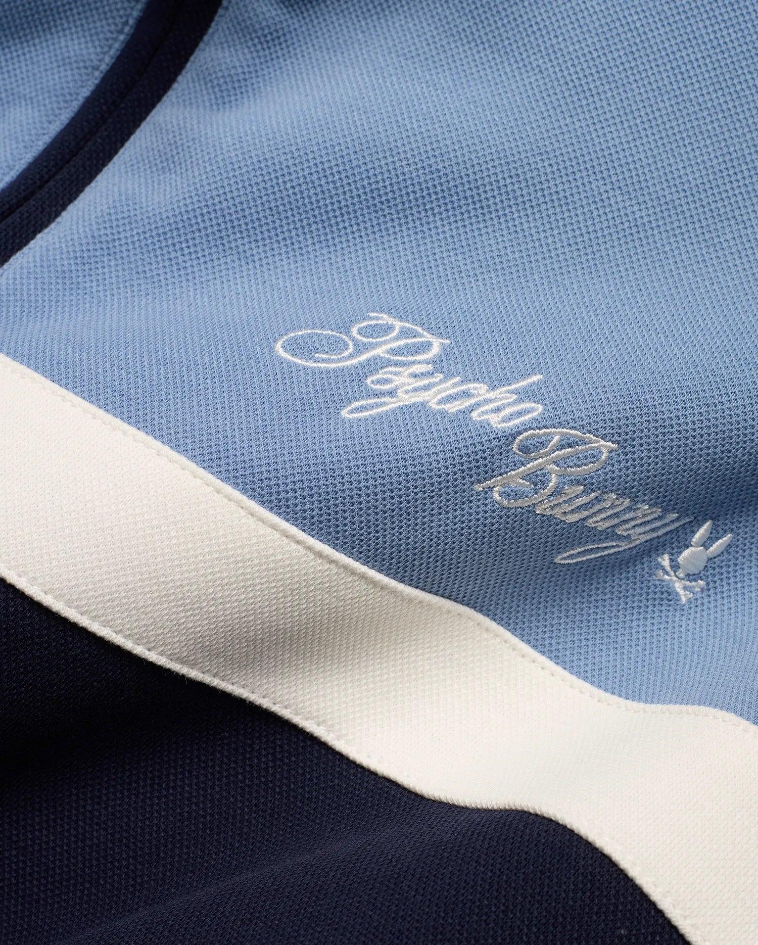 Men's Jacques Pique Track Jacket in light blue and navy pique fabric with a white stripe, featuring an embroidered white Psycho Bunny script logo and small bunny skull logo on the chest. (6)