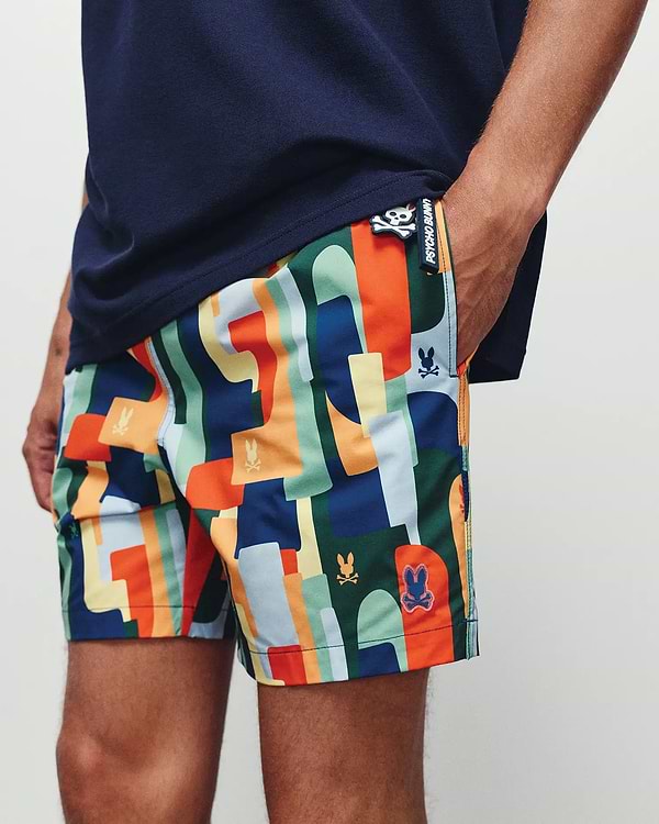 Mens Wes All Over Print Swim Trunk by Psycho Bunny, showcasing a vibrant multi-color abstract geometric pattern with integrated skull logos and a prominent branded side tag, classic fit.