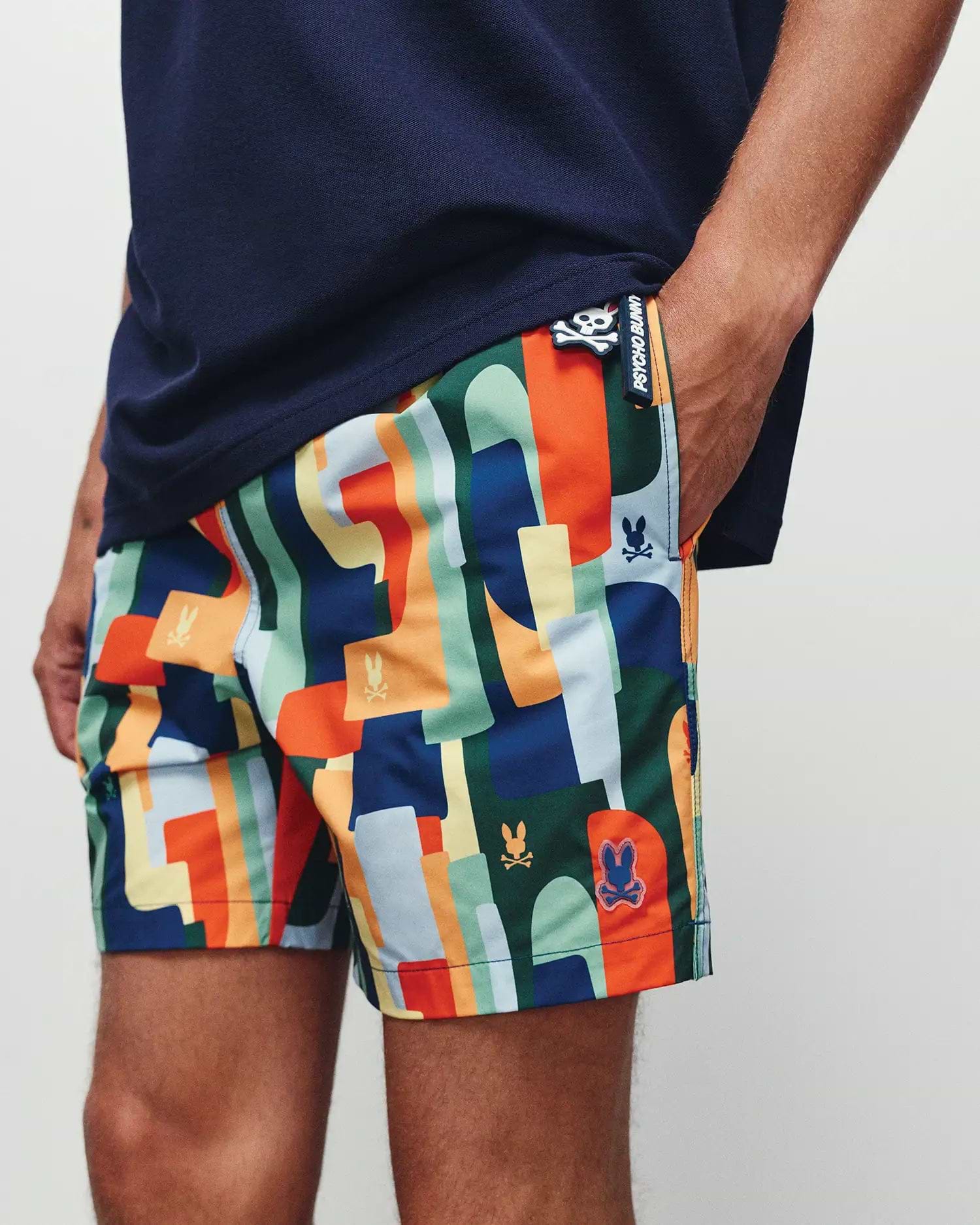 Mens Wes All Over Print Swim Trunk by Psycho Bunny, showcasing a vibrant multi-color abstract geometric pattern with integrated skull logos and a prominent branded side tag, classic fit.