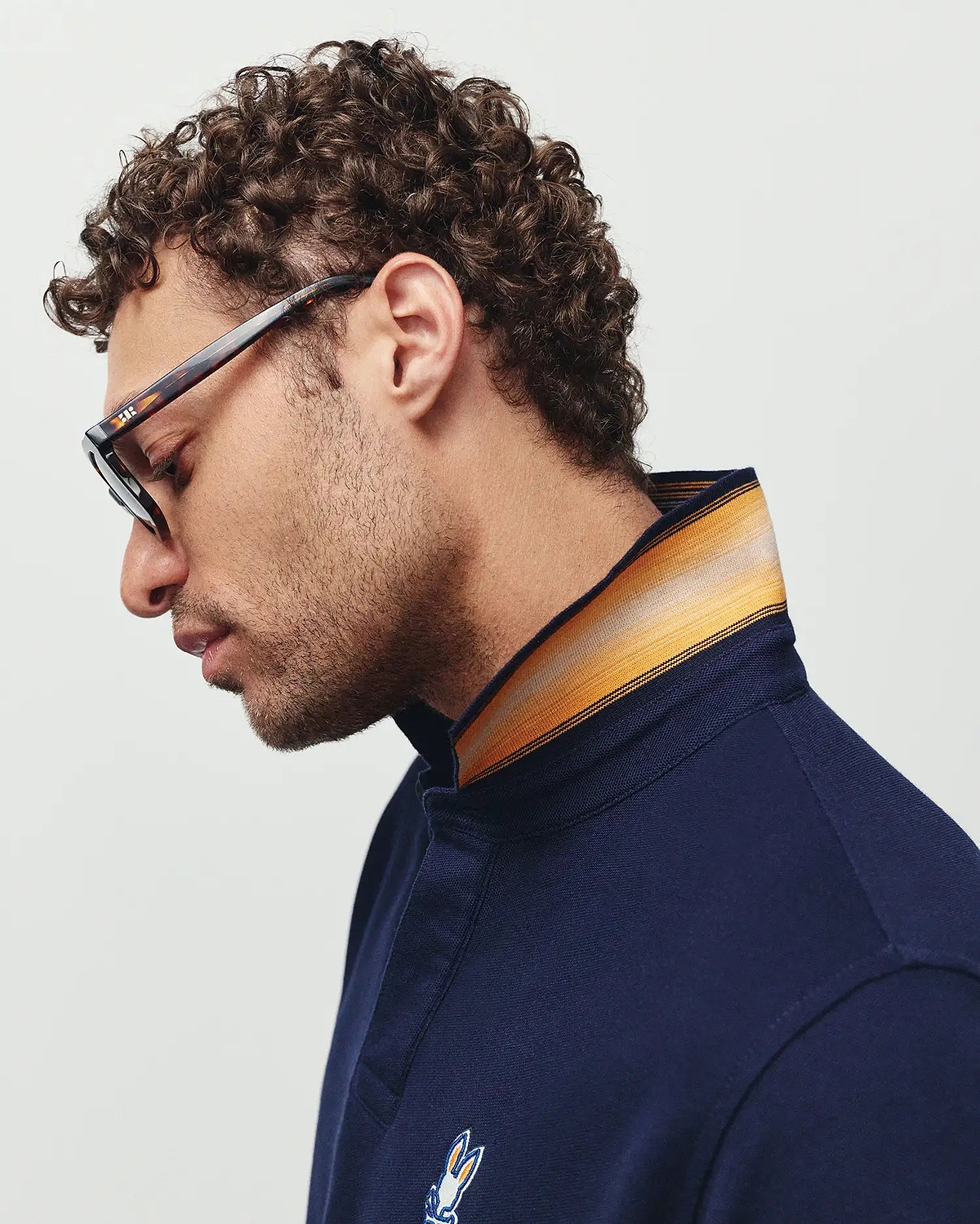 Mens Jaydon Pique Polo, a navy pique polo shirt with a classic fit, featuring a striped collar in orange, yellow, and grey, and a small embroidered Psycho Bunny skull logo on the left chest.