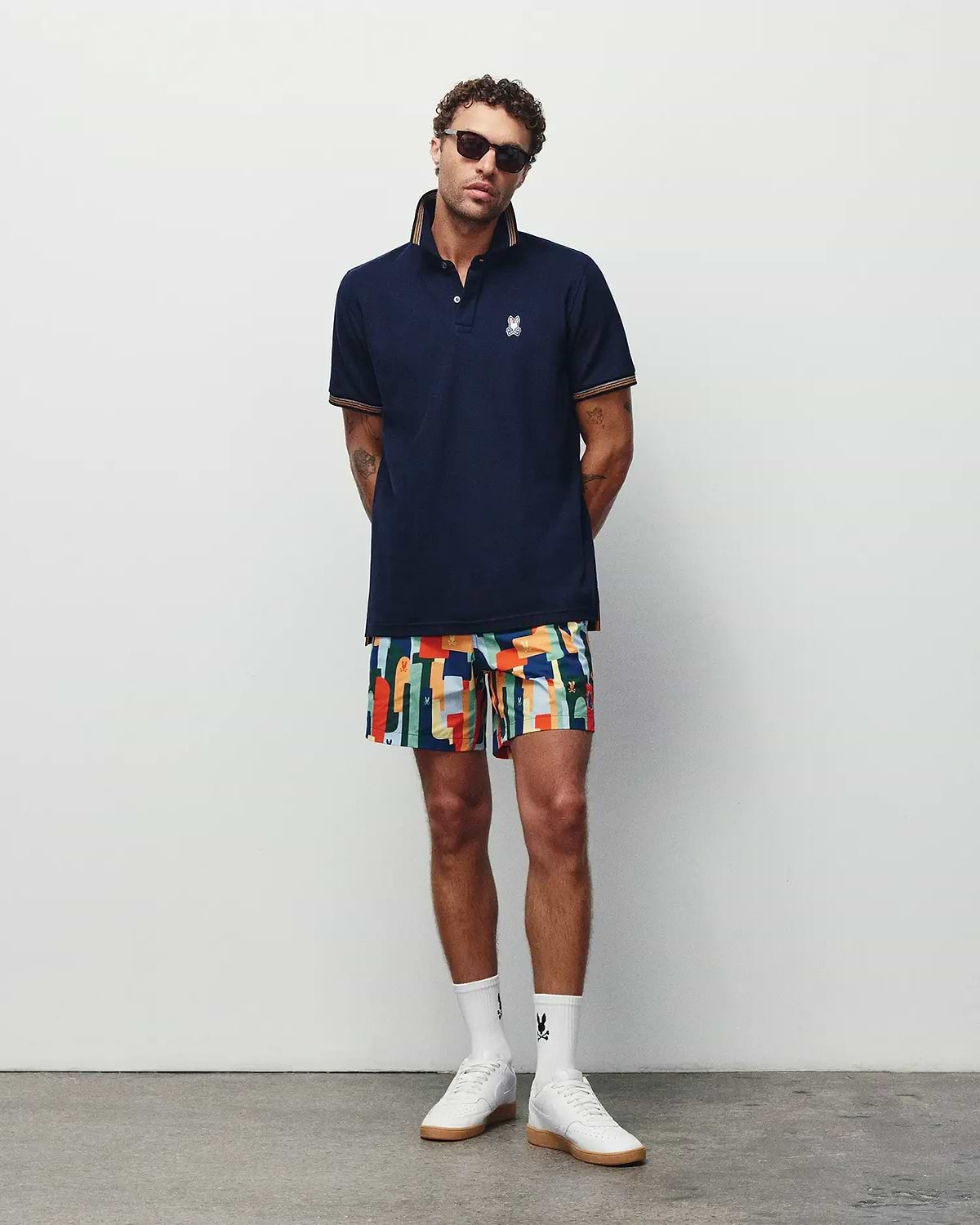 Navy pique polo with orange striped collar and cuffs, featuring a white embroidered Psycho Bunny logo on the chest, paired with vibrant Mens Wes All Over Print Swim Trunk shorts.
