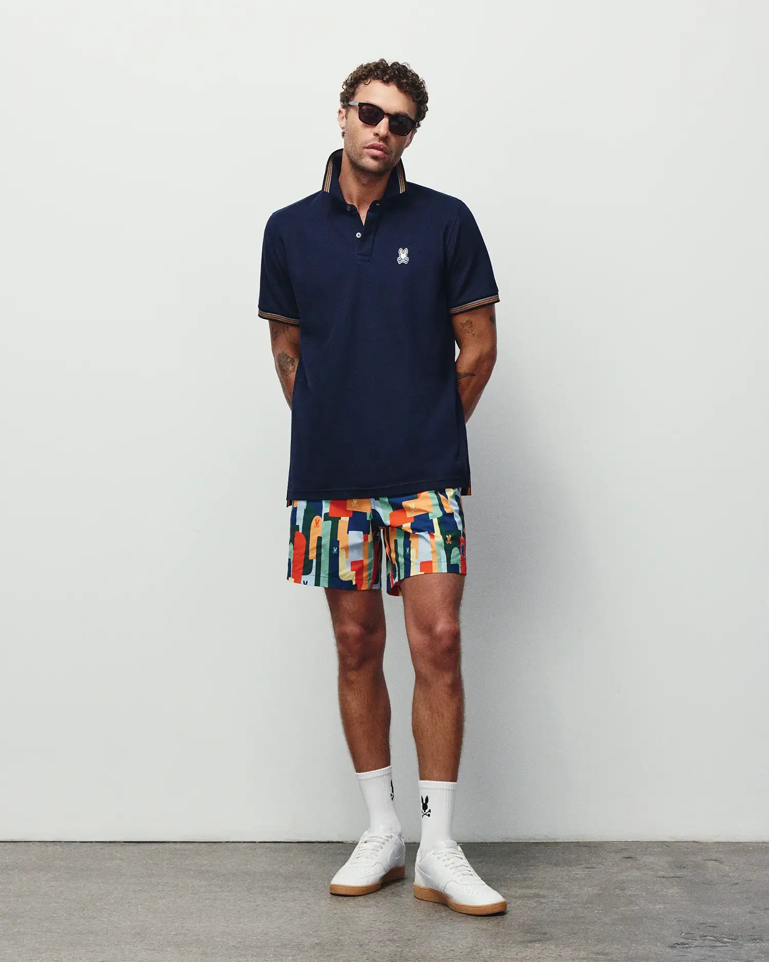 Navy pique polo with orange striped collar and cuffs, featuring a white embroidered Psycho Bunny logo on the chest, paired with vibrant Mens Wes All Over Print Swim Trunk shorts.