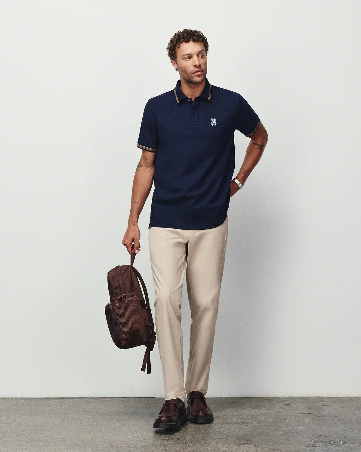 Navy pique polo with gold-striped collar and cuffs, featuring a small embroidered Psycho Bunny skull logo on the chest, paired with Mens Edward 10k Tapered Pant in light beige.