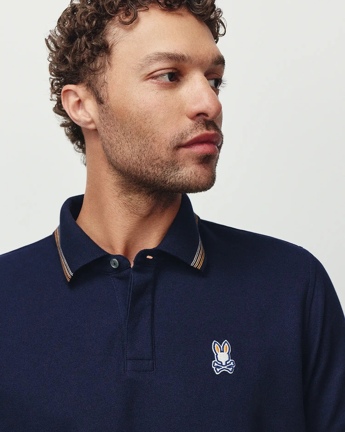 Mens Jaydon Pique Polo in navy blue, featuring a classic fit, textured pique fabric, a small embroidered Psycho Bunny skull logo on the left chest, and striped collar trim in orange, white, and silver.