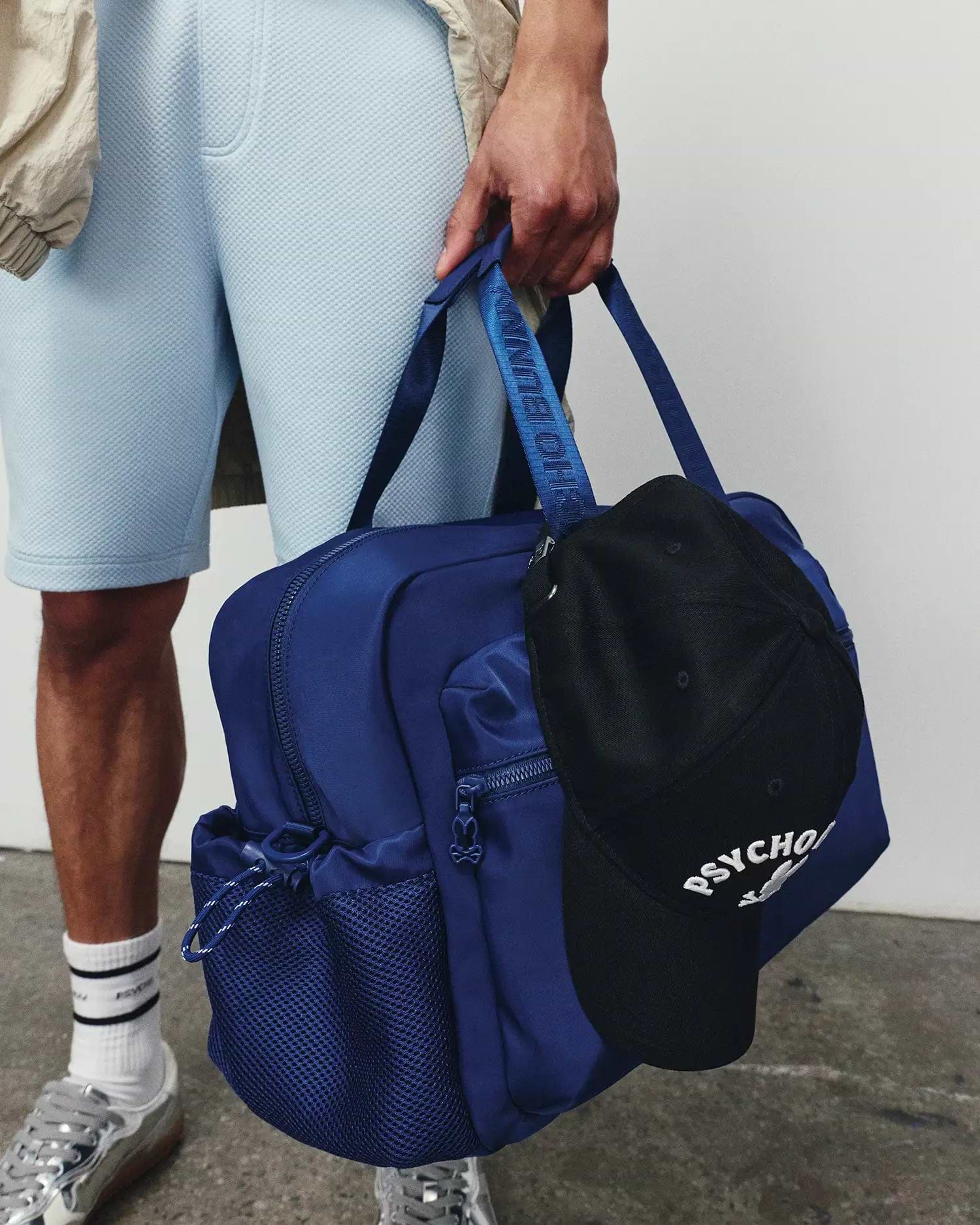 Light blue pique shorts, a beige jacket, and a royal blue backpack with a Psycho Bunny logo strap. A black Mens Ambrose Baseball Cap with white embroidered Psycho Bunny skull and text hangs from the bag.