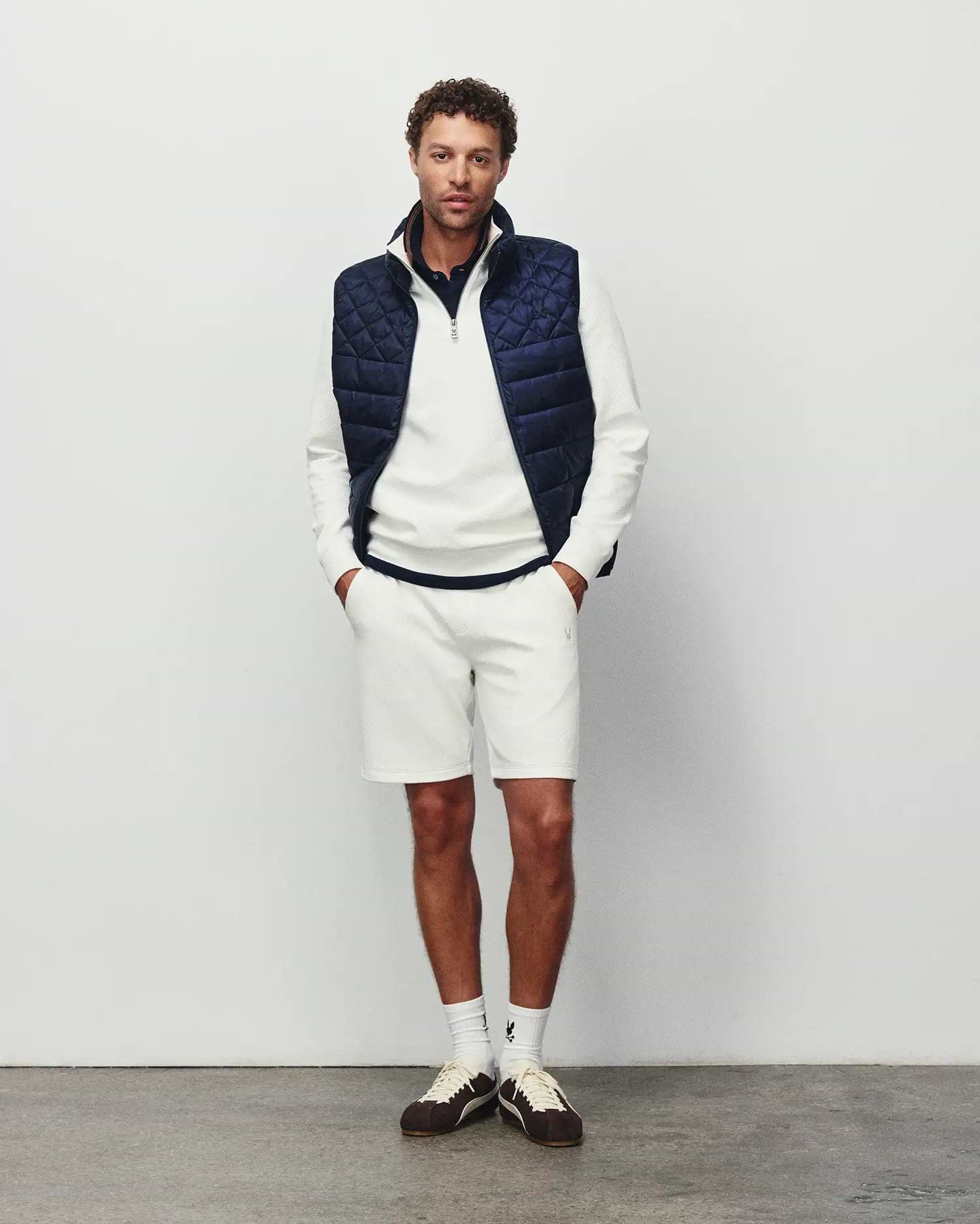 Mens Emerik Puffer Vest, navy blue, diamond quilted, with embroidered Psycho Bunny skull logo on chest. Worn with cream textured pique quarter-zip and matching shorts, both with bunny logo.