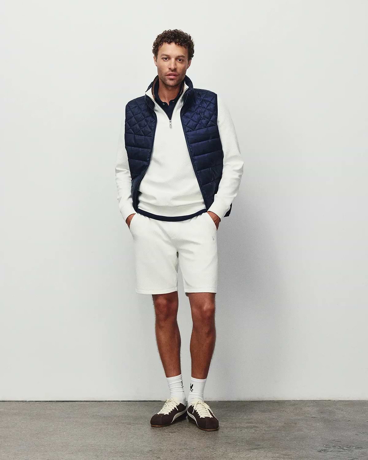 Mens Marc Textured Short in cream pique with a small embroidered Psycho Bunny logo on the left thigh, styled with a matching textured quarter-zip pullover featuring a chest logo and a navy quilted vest.