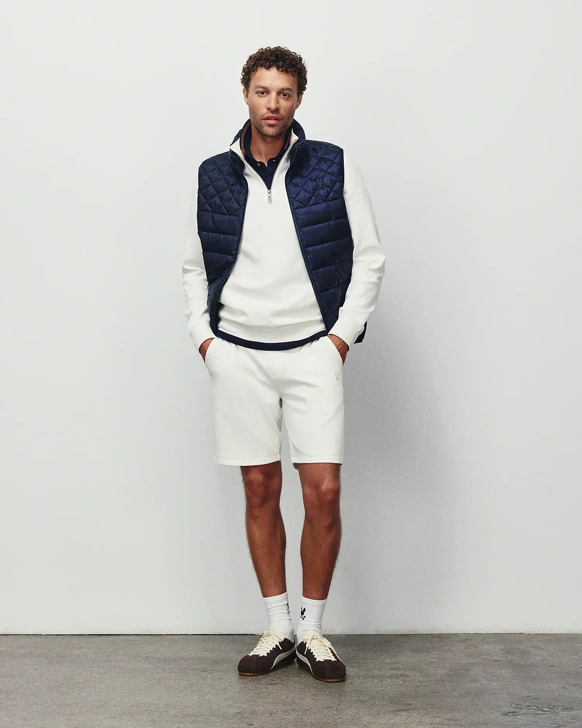 Mens Marc Textured Short in cream pique with a small embroidered Psycho Bunny logo on the left thigh, styled with a matching textured quarter-zip pullover featuring a chest logo and a navy quilted vest.