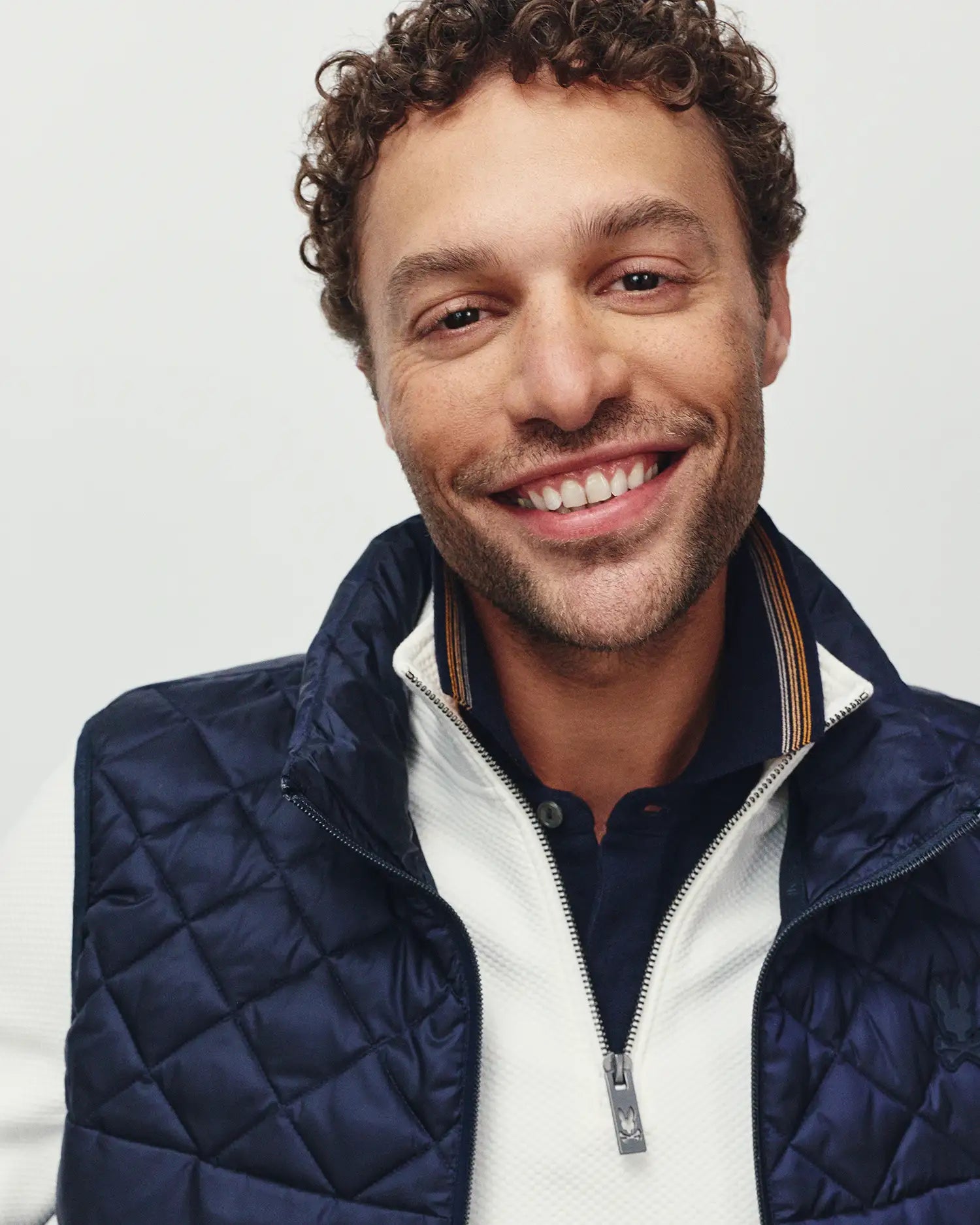 Mens Emerik Puffer Vest in navy with diamond quilting, featuring a small embroidered Psycho Bunny skull logo on the chest, layered over a white textured quarter-zip sweater with a logo zipper pull, and a navy polo with striped collar.