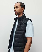 Mens Emerik Puffer Vest, a black quilted puffer with stand collar, full zip, and small embroidered Psycho Bunny skull logo on left chest, plus subtle text pattern on inner lining.