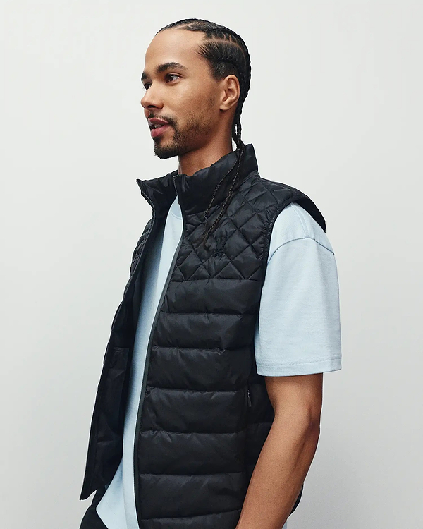 Mens Emerik Puffer Vest, a black quilted puffer with stand collar, full zip, and small embroidered Psycho Bunny skull logo on left chest, plus subtle text pattern on inner lining.