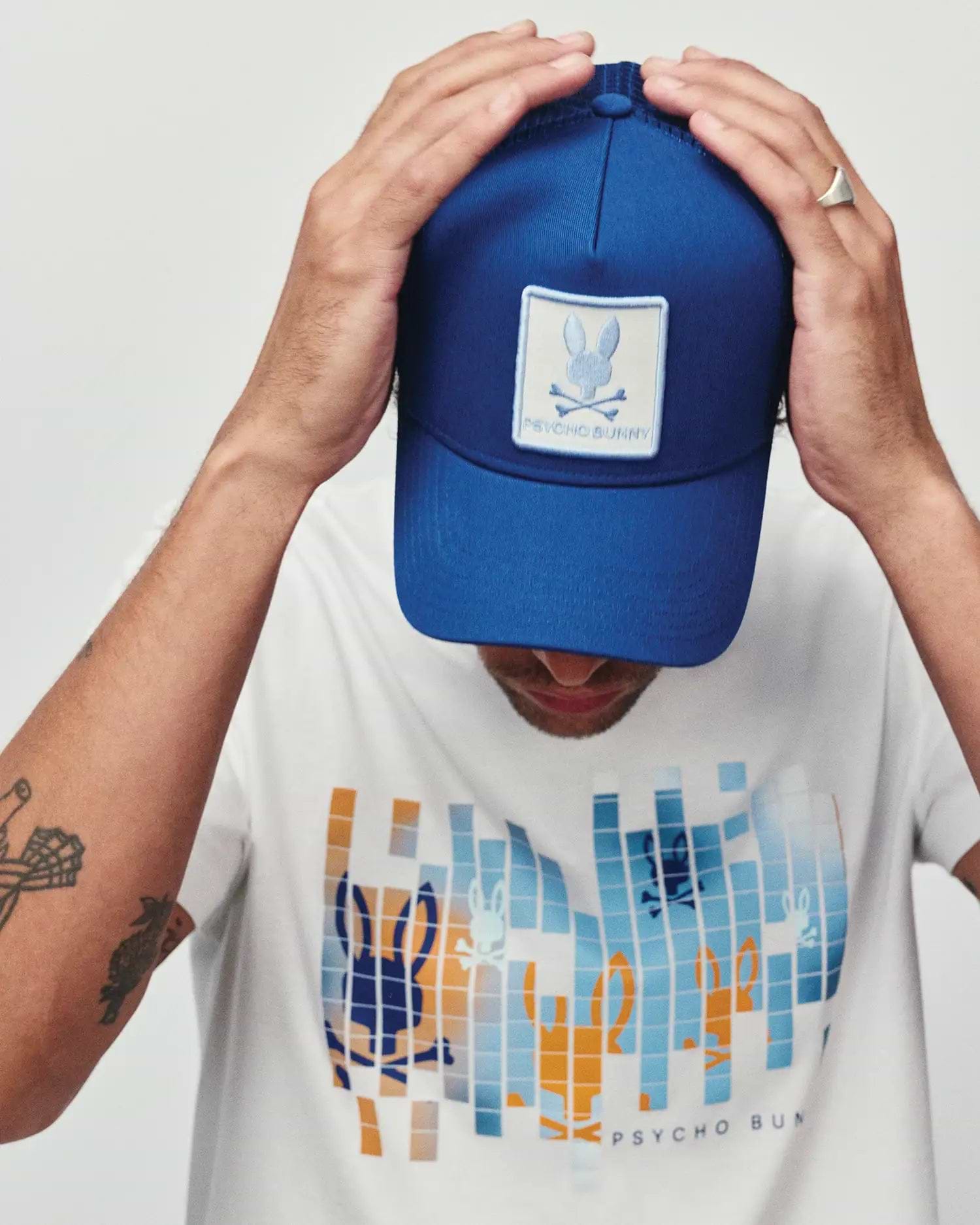 Mens Adamo Bunny Trucker Cap in royal blue with a white embroidered Psycho Bunny skull patch, paired with a white classic fit jersey t-shirt featuring a large pixelated blue and orange Psycho Bunny graphic print on the chest.