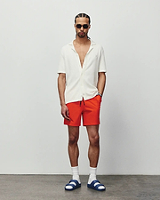 Mens Samos Hydrochromic Printed Swim Trunk in vibrant orange, classic fit, paired with a relaxed white ribbed knit short-sleeve collared shirt featuring a small embroidered Psycho Bunny logo on the left chest.