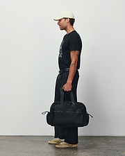 Black classic fit jersey t-shirt with a large white Psycho Bunny skull graphic, relaxed black pants, a cream cap, and a black Mens Nylon Duffle Bag featuring a subtle logo. (2)