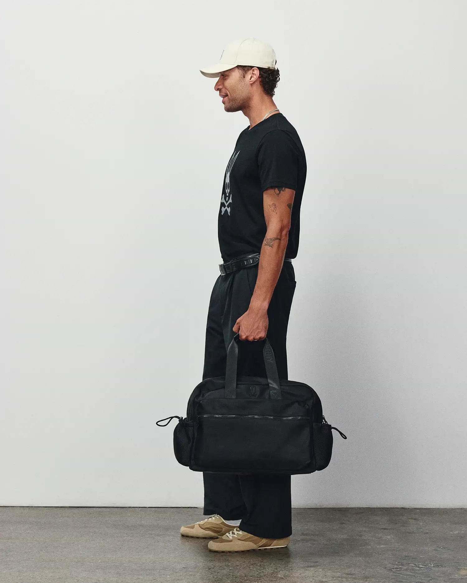 Black classic fit jersey t-shirt with a large white Psycho Bunny skull graphic, relaxed black pants, a cream cap, and a black Mens Nylon Duffle Bag featuring a subtle logo. (2)