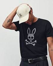 MENS STAN GRAPHIC TEE in black jersey, featuring a large white checkered Psycho Bunny skull and crossbones graphic on the chest. Accessorized with a cream cap with a small embroidered pink Psycho Bunny logo and a black leather belt.
