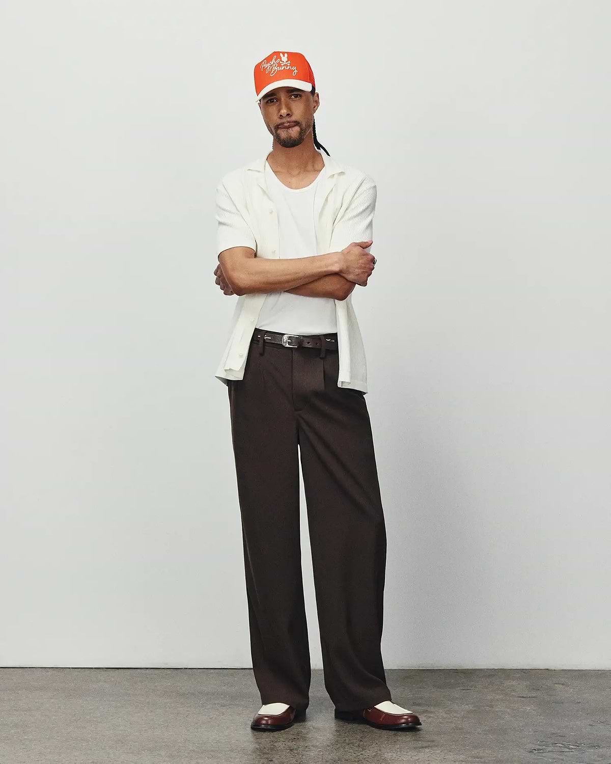 Mens Yann Knit Shirt: Relaxed fit, off-white ribbed knit short-sleeve collared shirt, layered over a white tee, with dark brown wide-leg trousers and an orange Psycho Bunny embroidered logo cap.
