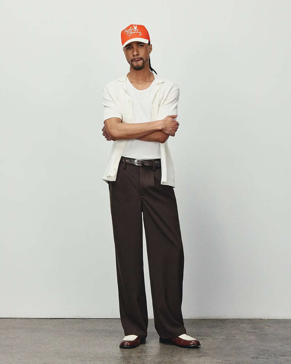 Mens Yann Knit Shirt: Relaxed fit, off-white ribbed knit short-sleeve collared shirt, layered over a white tee, with dark brown wide-leg trousers and an orange Psycho Bunny embroidered logo cap.