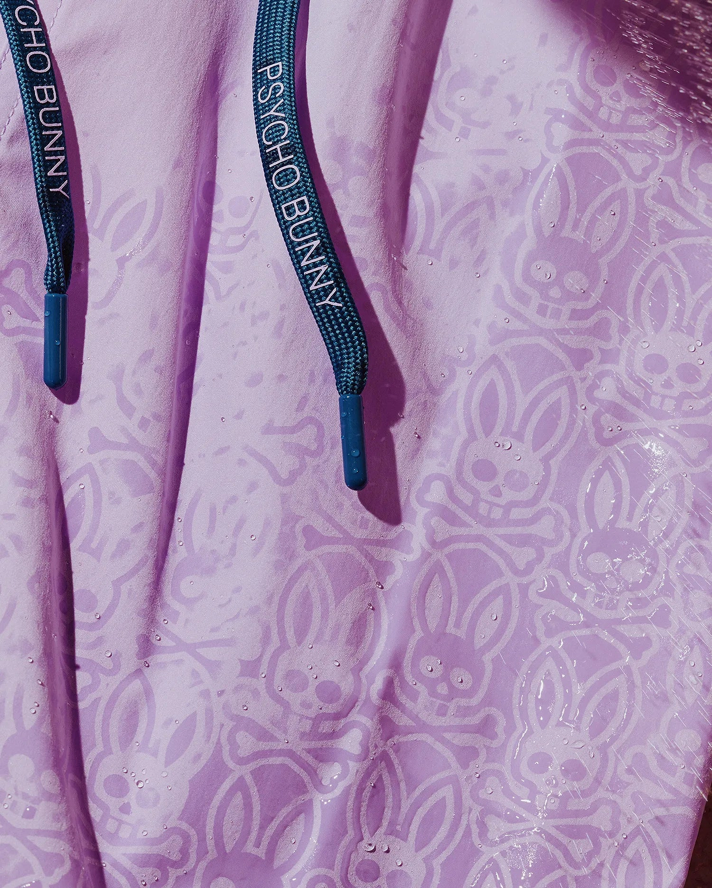 Mens Samos Hydrochromic Printed Swim Trunk in lavender, featuring an all-over subtle bunny skull and crossbones hydrochromic print, visible with water droplets. Dark teal drawstrings with Psycho Bunny text and blue tips.