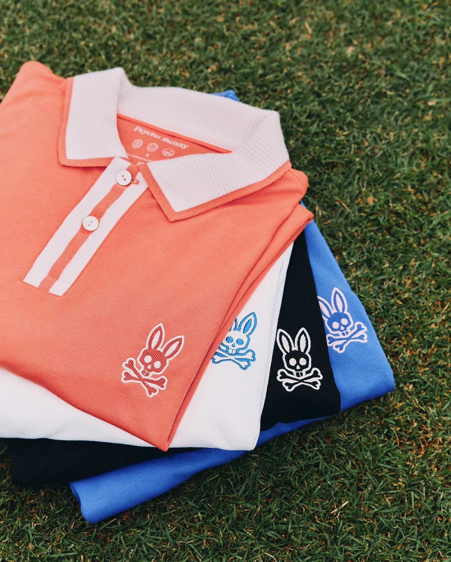 Mens Ridley Pique Polo stack: coral with white ribbed collar and placket stripes, white, black, and royal blue pique polos, each featuring a small embroidered Psycho Bunny skull logo on the left chest.