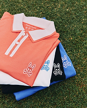 Mens Ridley Pique Polo stack: coral with white ribbed collar and placket stripes, white, black, and royal blue pique polos, each featuring a small embroidered Psycho Bunny skull logo on the left chest.