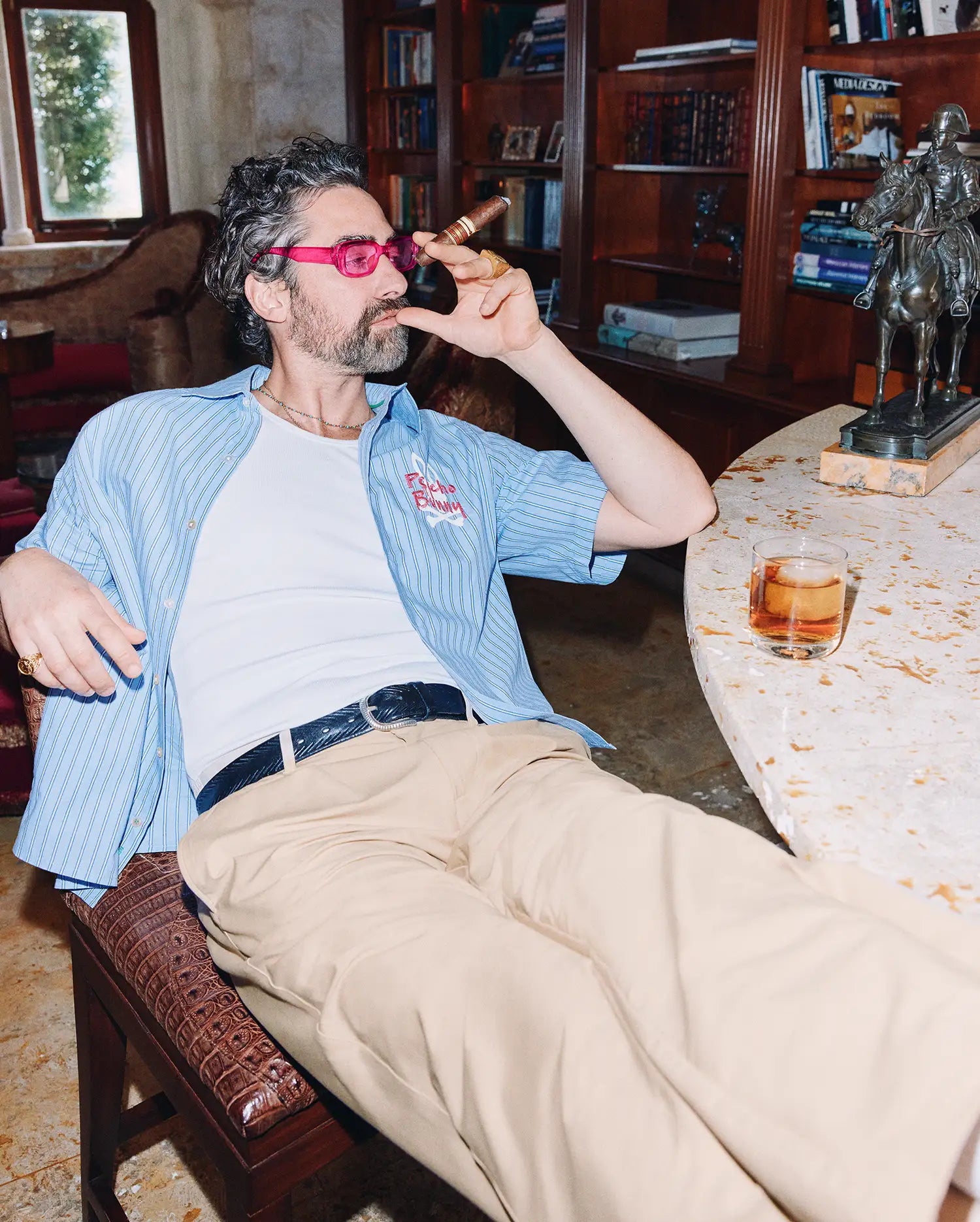 Mens Duncan Stripe Shirt, a light blue short-sleeve button-up with thin white stripes, relaxed fit, and a pink embroidered Psycho Bunny logo on the chest, paired with a white ribbed t-shirt and khaki trousers. (1)