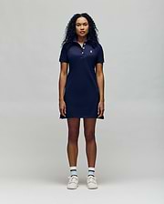 A woman with curly dark hair stands facing forward in a navy blue Psycho Bunny WOMENS POLO DRESS - WO2000003, paired with white socks with blue stripes and white sneakers, set against a plain light gray background.