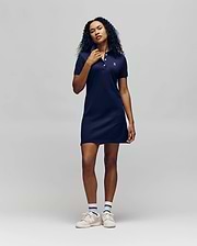 A person with long curly hair wears the Psycho Bunny WOMENS POLO DRESS - WO2000003 in navy blue, paired with white sneakers and striped socks, posing against a plain light grey background.