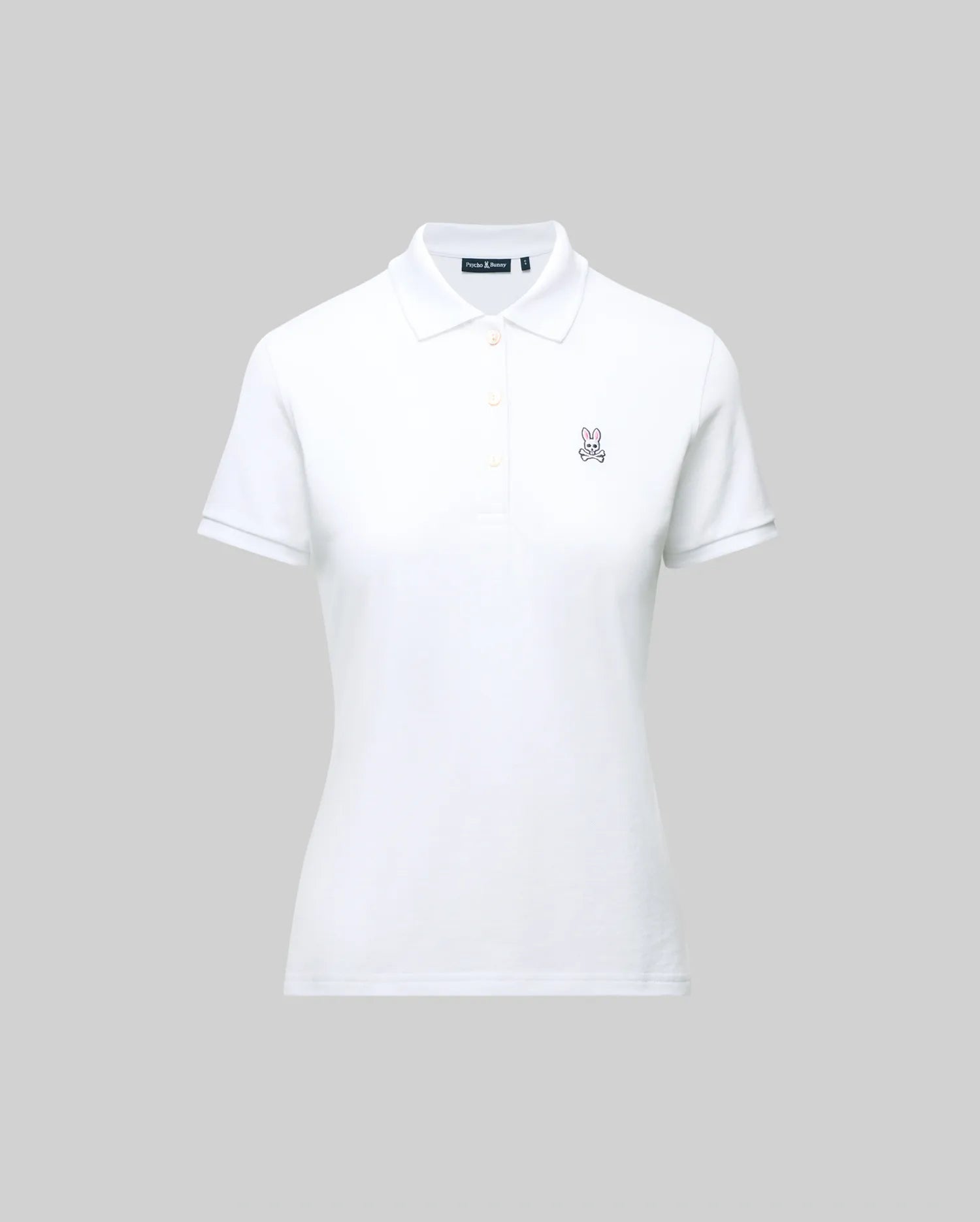 The WOMENS PIQUE POLO - WT0100003 by Psycho Bunny is a white short-sleeve polo featuring a collar, button placket, and 4k-stitch embroidered Bunny logo on the left chest, shown on a plain gray background.