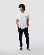 Against a minimal backdrop, an individual with curly hair is styled in a Psycho Bunny's white MENS CHICAGO CITY TEE - B6U551W1PC, featuring a small, vibrant Bunny logo graphic. The outfit is enhanced with black pants and black sneakers with contrasting white soles, made from soft Pima cotton jersey. The person nonchalantly has their hands in their pockets.