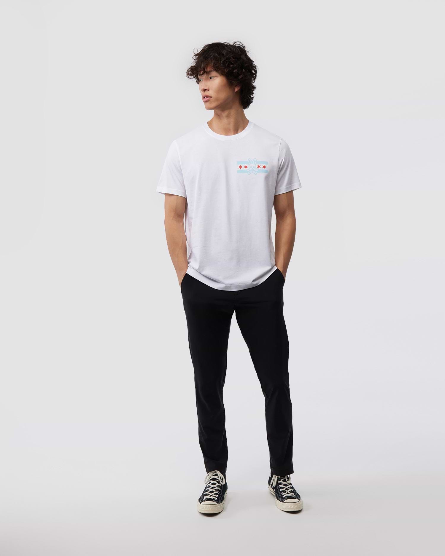 Against a minimal backdrop, an individual with curly hair is styled in a Psycho Bunny's white MENS CHICAGO CITY TEE - B6U551W1PC, featuring a small, vibrant Bunny logo graphic. The outfit is enhanced with black pants and black sneakers with contrasting white soles, made from soft Pima cotton jersey. The person nonchalantly has their hands in their pockets.