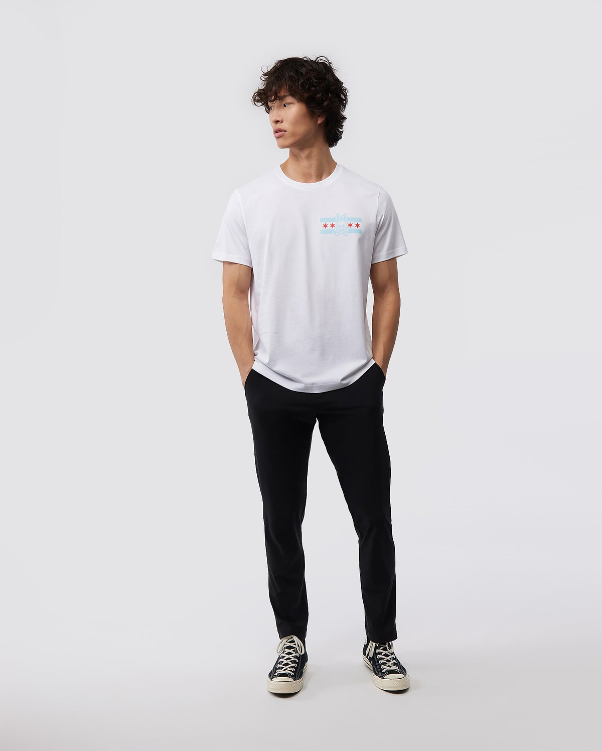 Against a minimal backdrop, an individual with curly hair is styled in a Psycho Bunny's white MENS CHICAGO CITY TEE - B6U551W1PC, featuring a small, vibrant Bunny logo graphic. The outfit is enhanced with black pants and black sneakers with contrasting white soles, made from soft Pima cotton jersey. The person nonchalantly has their hands in their pockets.