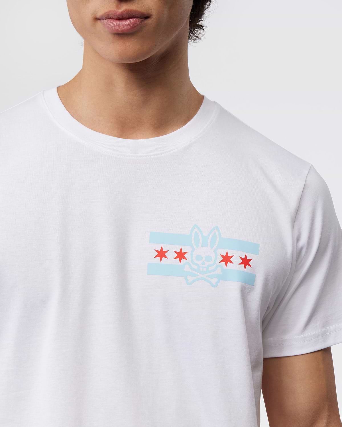A person wears the Psycho Bunny Men's Chicago City Tee - B6U551W1PC, crafted from soft Pima cotton jersey and featuring a bunny logo graphic. The design includes a stylized bunny skull and crossbones, two light blue horizontal stripes, and five red stars on the chest against a plain background.