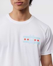 A person wears the Psycho Bunny Men's Chicago City Tee - B6U551W1PC, crafted from soft Pima cotton jersey and featuring a bunny logo graphic. The design includes a stylized bunny skull and crossbones, two light blue horizontal stripes, and five red stars on the chest against a plain background.