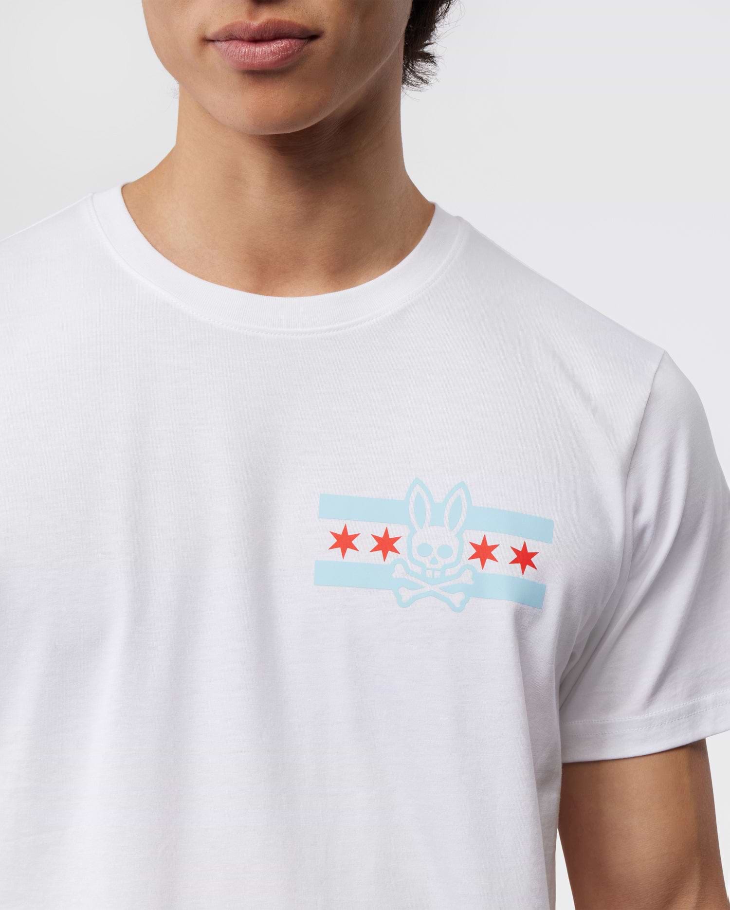 A person wears the Psycho Bunny Men's Chicago City Tee - B6U551W1PC, crafted from soft Pima cotton jersey and featuring a bunny logo graphic. The design includes a stylized bunny skull and crossbones, two light blue horizontal stripes, and five red stars on the chest against a plain background.
