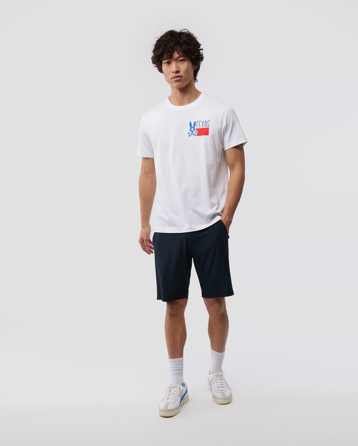 A young man with curly hair stands against a plain background, wearing a white MENS TEXAS TEE - B6U553W1PC featuring the Psycho Bunny logo graphic, navy shorts, white socks, and sneakers.